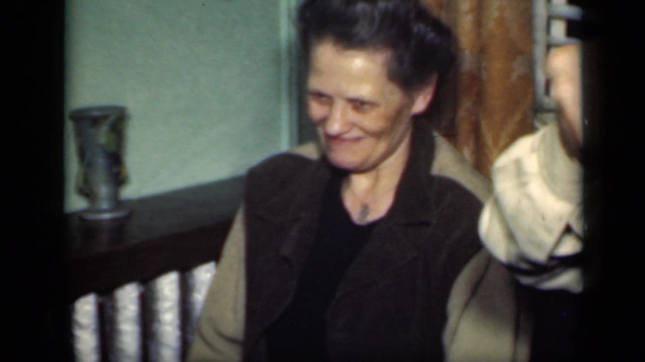 Woman in dark vest looks down and smiles, with another person partly visible beside her.