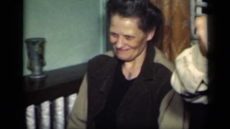1949 New York home-movie frame with an adult woman looking down as she smiles, seated close to another partly visible adult indoors.