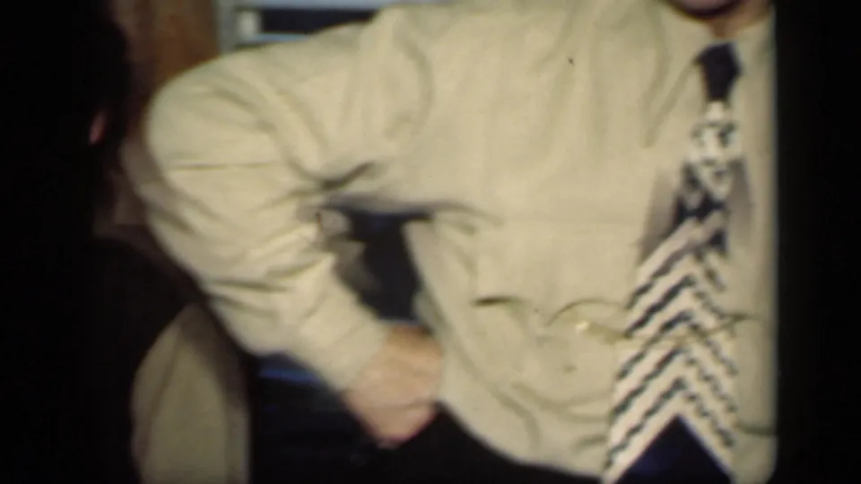 1949 New York home-movie frame showing a cropped seated adult man in a light shirt and bold patterned tie inside a private interior.