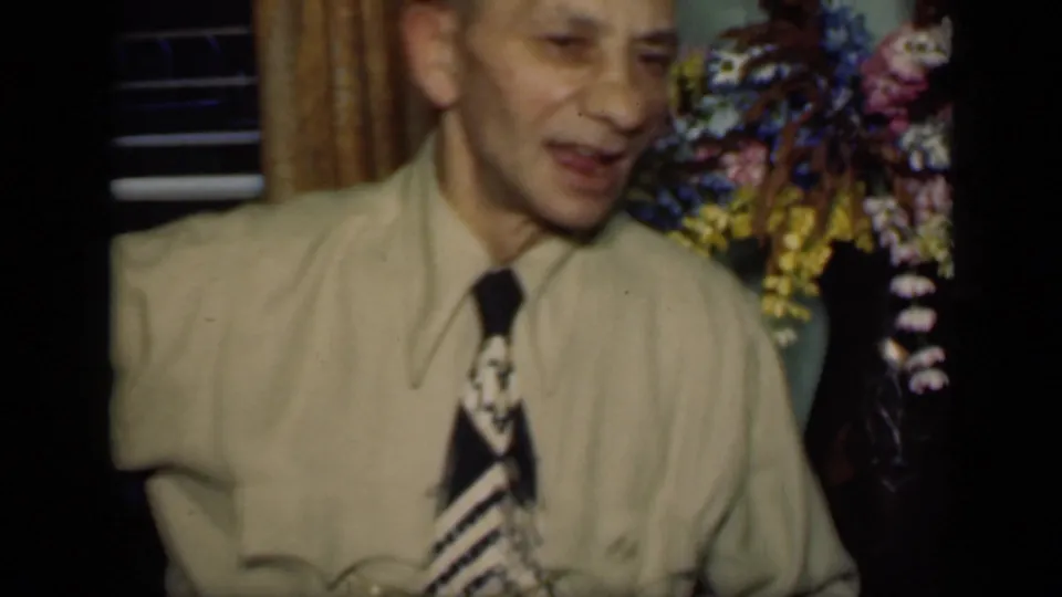 1949 New York home-movie frame of an adult man in a light shirt and patterned tie seated beside flowers in a domestic interior.