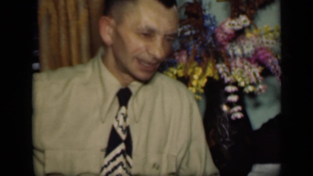 Adult man in a light shirt and patterned tie sits near flowers in a dim interior.