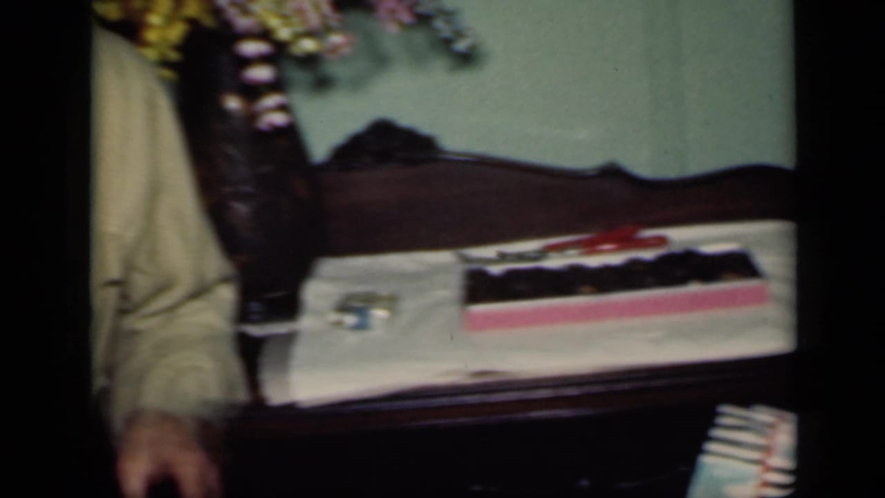 Box of candy rests on a bed or table surface beside flowers and a partial figure.