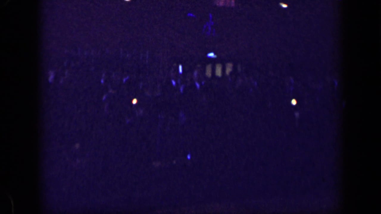 Dark indoor crowd with blue-purple light and small bright points in the distance.