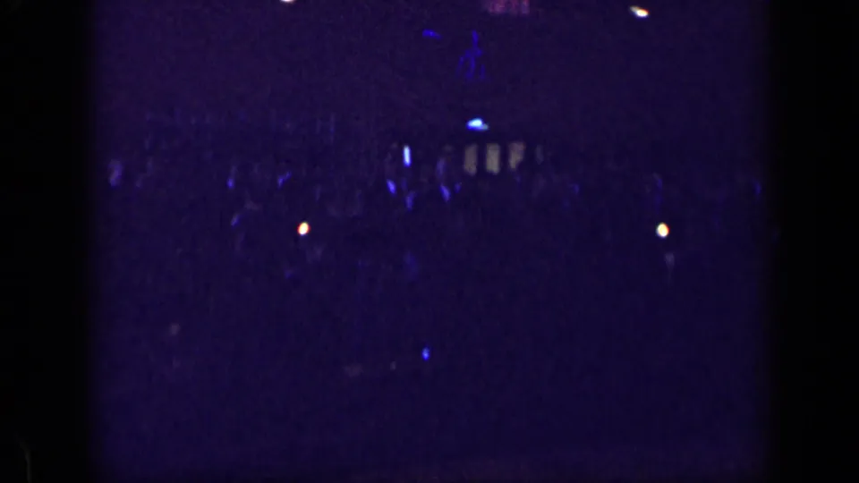 1957 New York 8mm frame showing a dim indoor crowd under blue-purple light, with people mostly reduced to silhouettes.