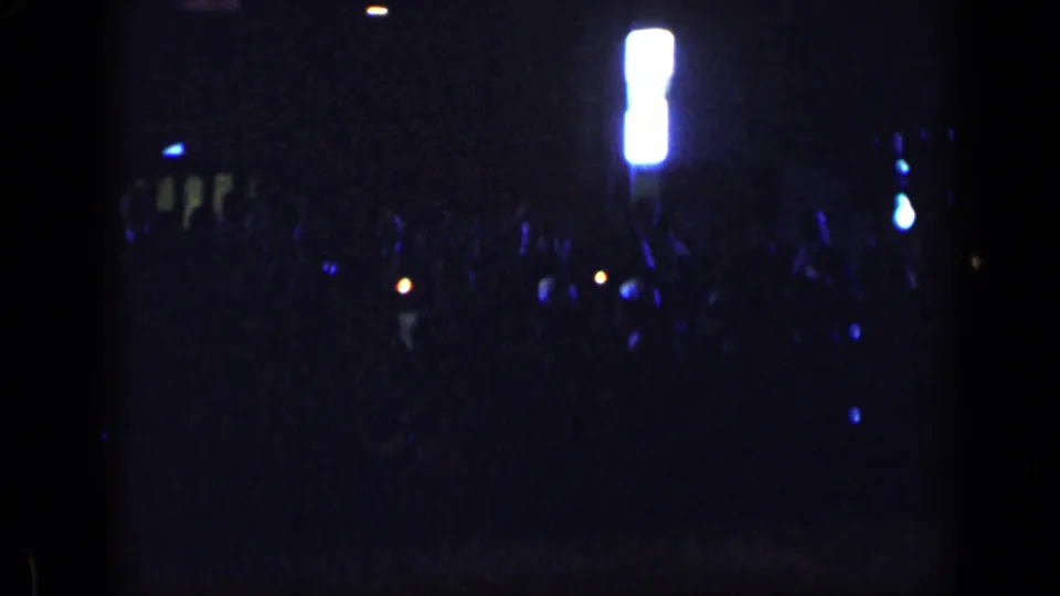 1957 New York 8mm footage frame with a dark indoor crowd, a bright white vertical fixture, and scattered blue and orange lights.