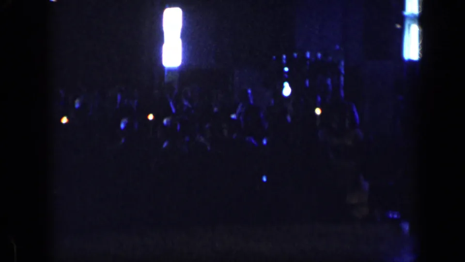 Late frame from a 1957 New York 8mm clip showing a dim crowd, bright vertical lights, and blue reflections in a dark interior.