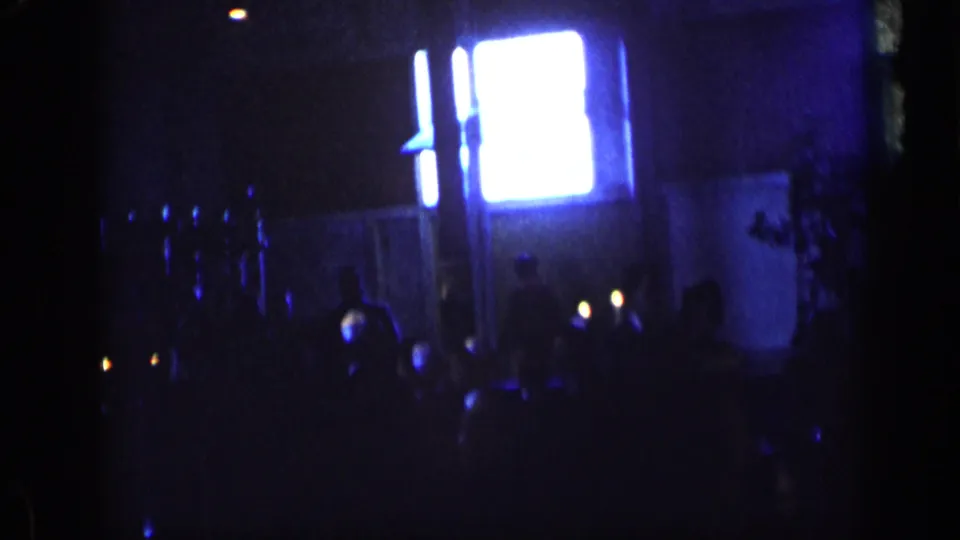 1957 New York home-movie frame with a bright rectangular light source above indistinct pedestrians and dark blue street shadows.