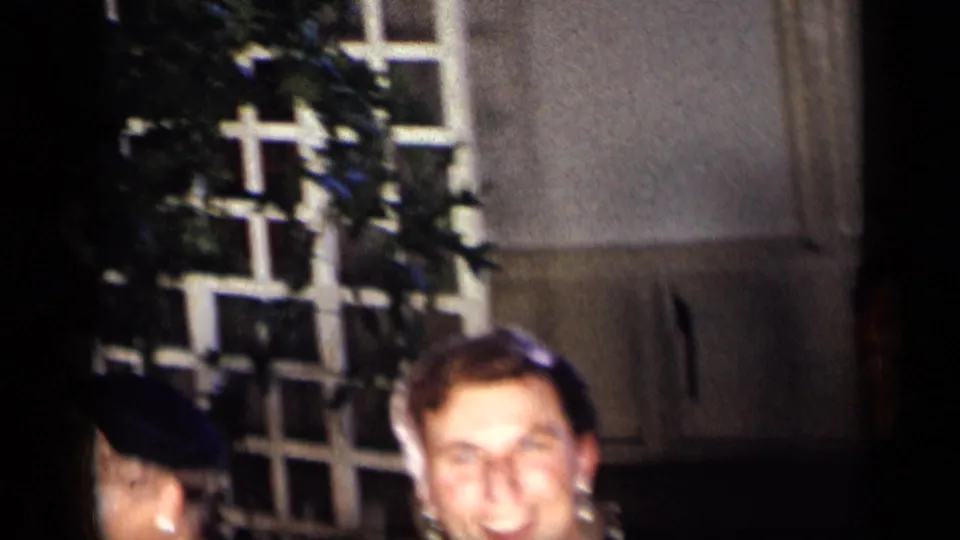 1957 New York dinner-party frame showing cropped adult faces near a white lattice trellis and leafy greenery in the background.