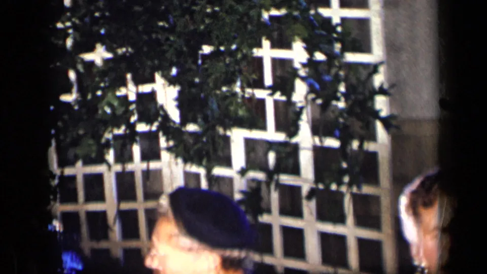 1957 New York home-movie frame with white lattice, leafy plants, and partial adult guests seated below the camera line.