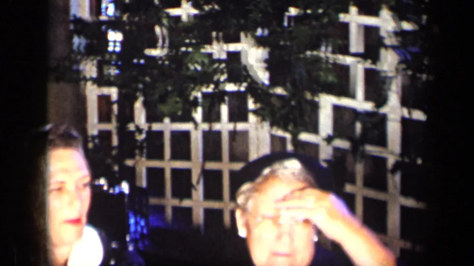 1957 New York dinner-party frame of seated adults beneath a white lattice trellis, with one guest lifting a hand toward their face.