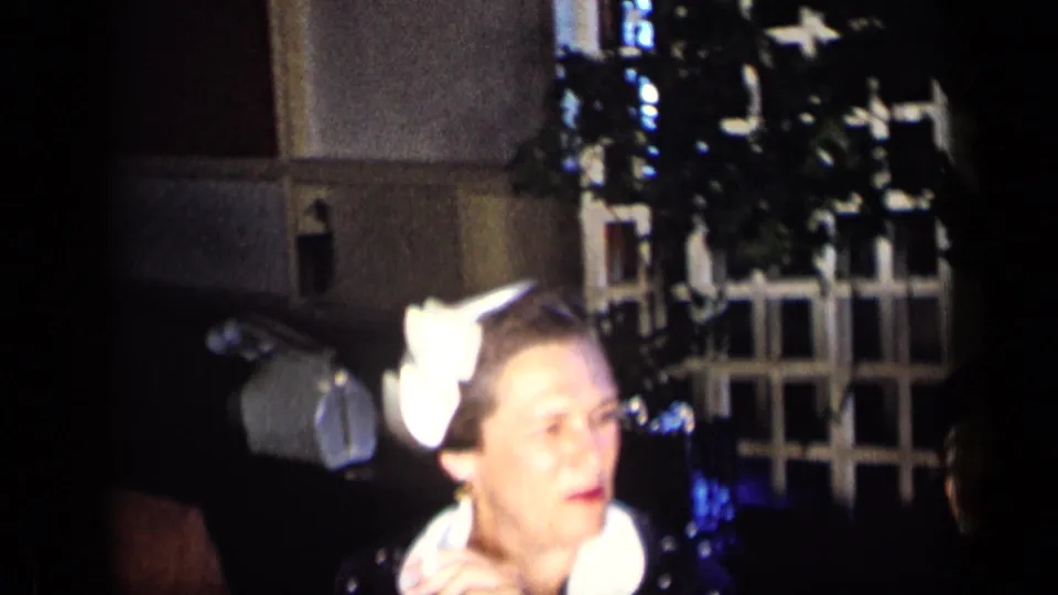 1957 New York home-movie frame showing a woman with a white hair bow and a suited man in a dark party interior.