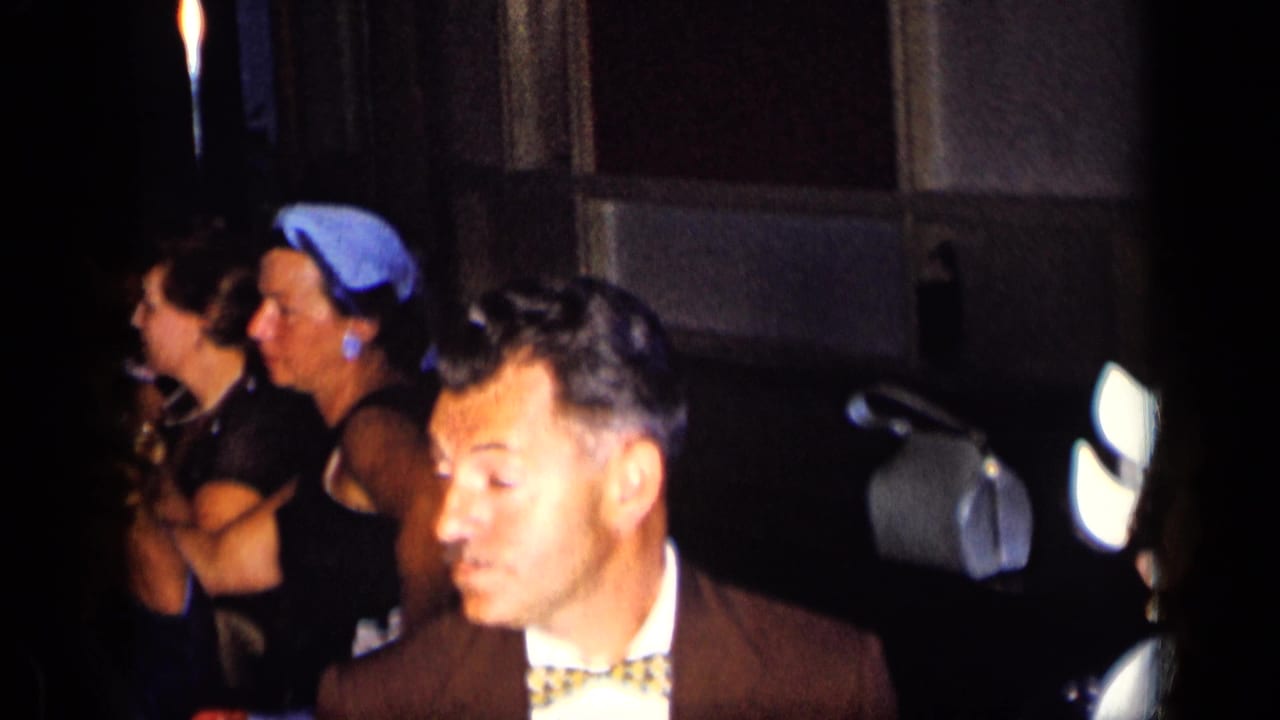 1957 New York party frame with formally dressed adults, including a woman in a blue headscarf, seen under warm low light.