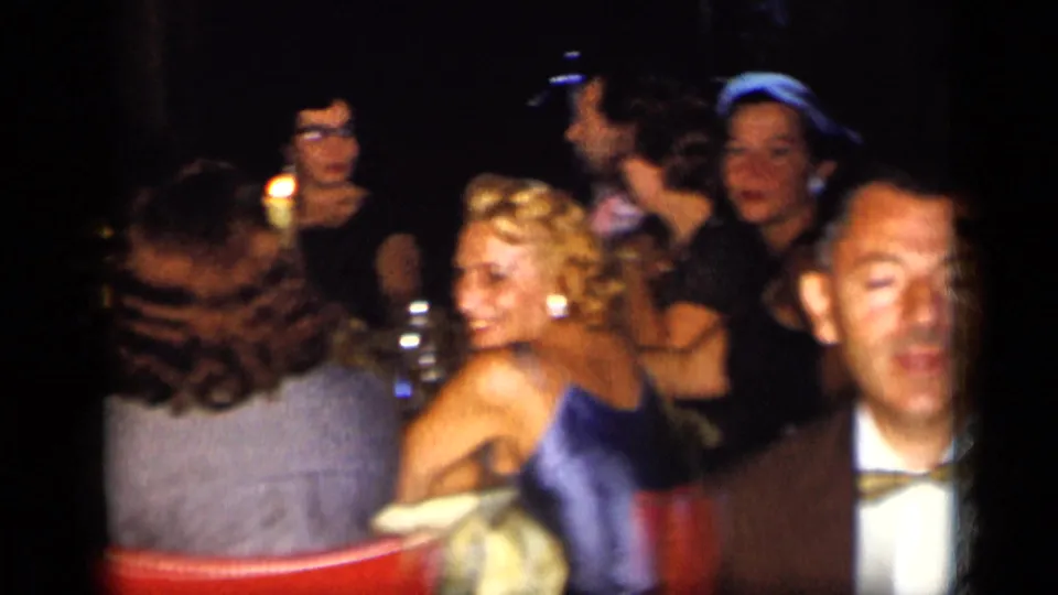 1957 New York home-movie frame of adults seated at a formal party table with red chairs and low candlelit atmosphere.