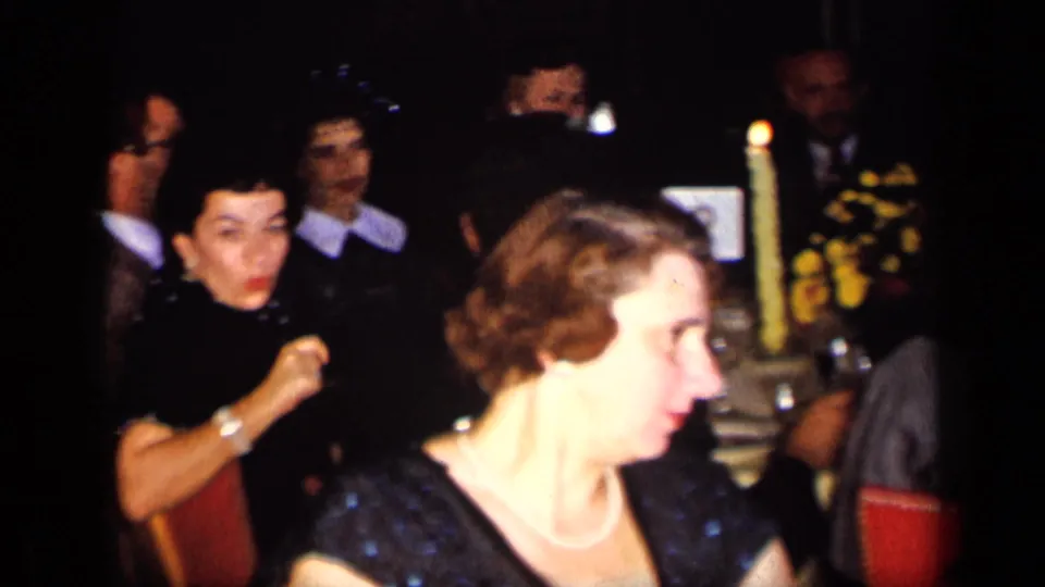 1957 New York party frame showing seated adults at a table, including a woman in a dark dress and visible bottles nearby.