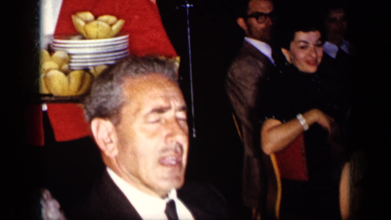 Man in tuxedo appears near a waiter with bread trays and guests in evening wear.