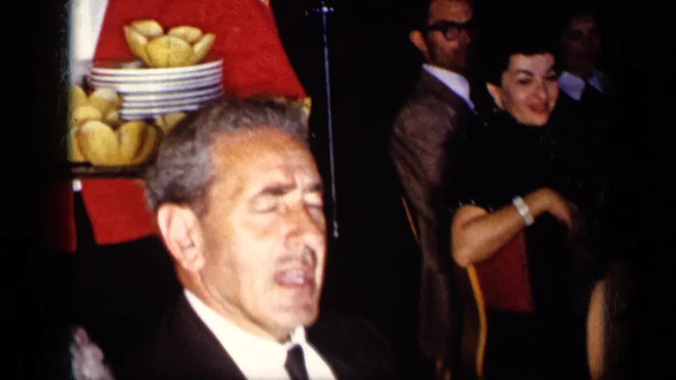 1957 New York formal banquet view with a tuxedoed man, red-jacketed waiter, bread trays, and guests in dark evening clothing.