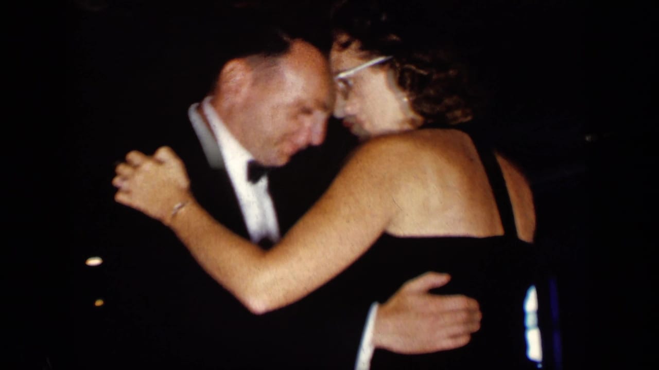 Man in a tuxedo dances closely with a woman in a dark dress in a dim room.