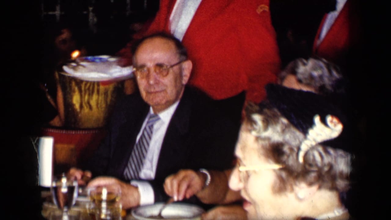Seated adults at a candlelit restaurant table with a waiter in a red jacket nearby.
