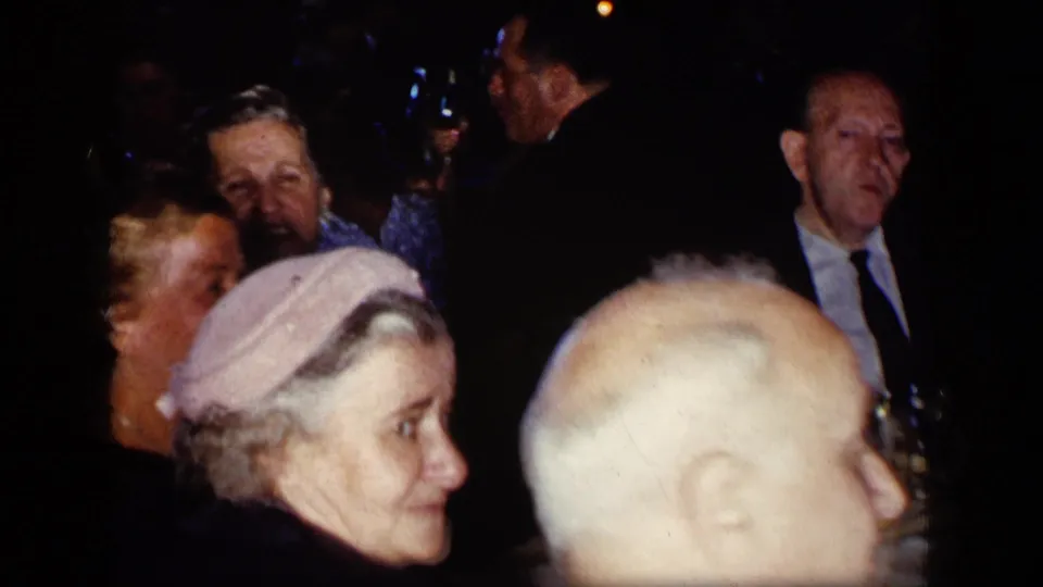 Well-dressed adults in a dim 1957 New York restaurant setting, including a woman in a pale hat and men in suits at close tables.