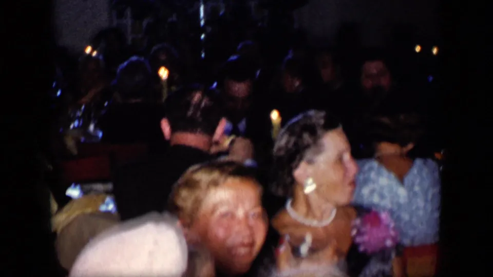 A crowded 1957 New York restaurant dinner, filmed from within the room, with seated adults, candles, dresses, and suits visible.