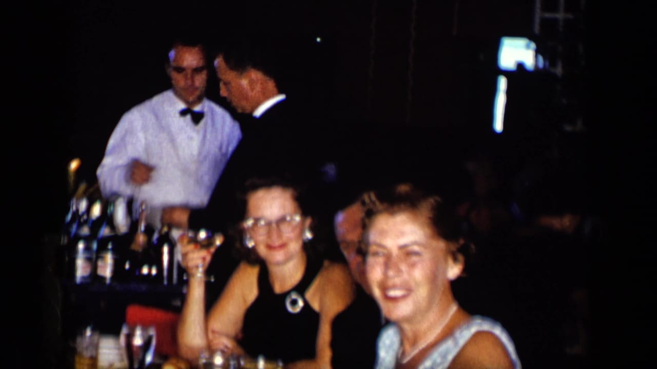 Adults seated at a dim restaurant table while a waiter stands near bottles behind them.