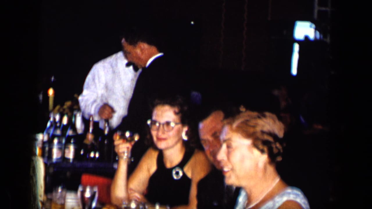 Two women sit with drinks at a table while a waiter and another man stand nearby.