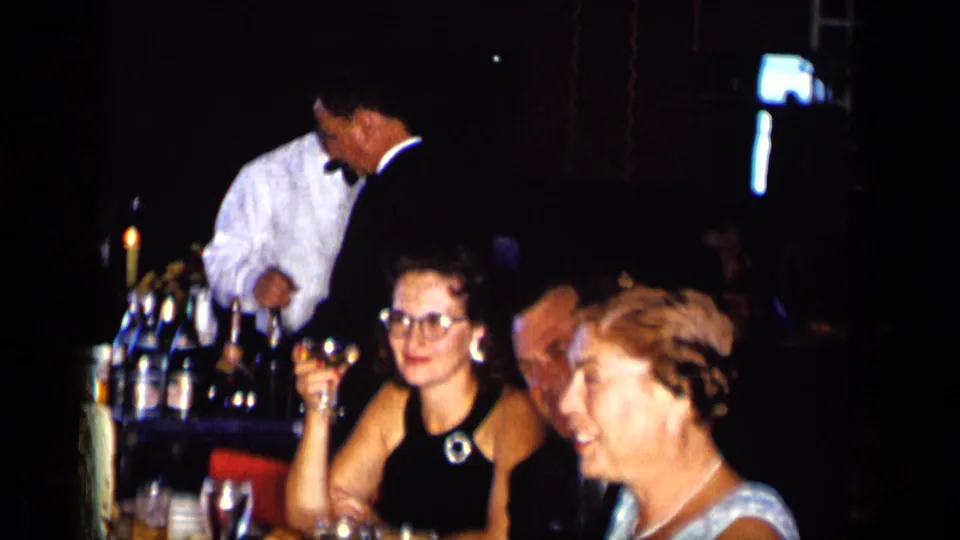 Adults in formal attire sit at a dim New York restaurant or bar table in 1957, with drinks, bottles, and a white-jacketed waiter visible.