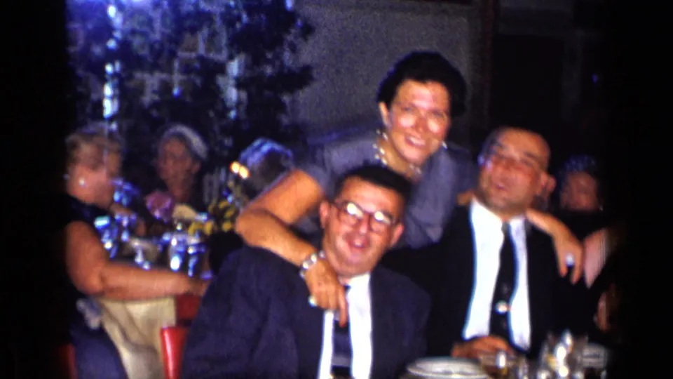Three adults smile and lean together for the 1957 New York home-movie camera at a table with drinks in a dark restaurant setting.