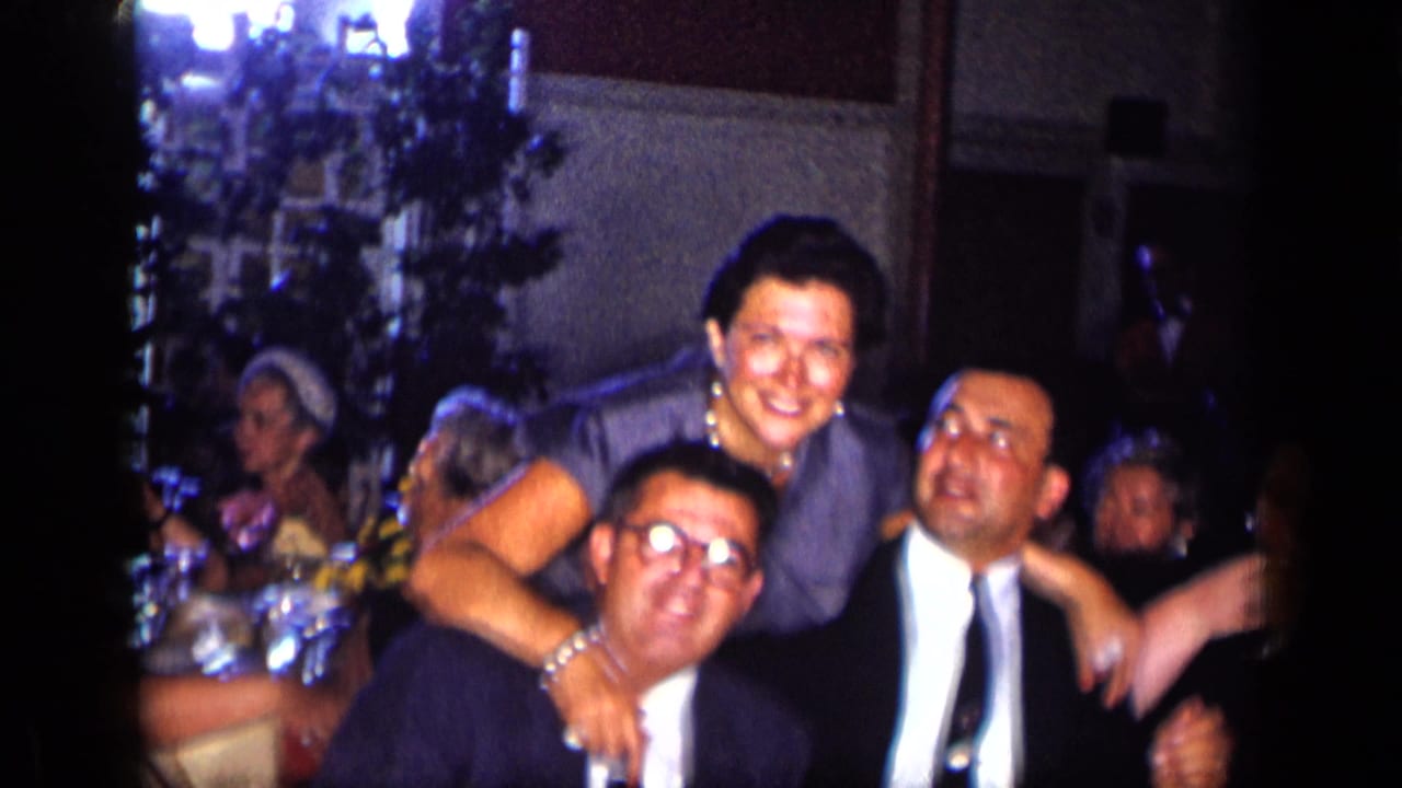 Adults seated at a dinner table, with a woman leaning over two men in suits.