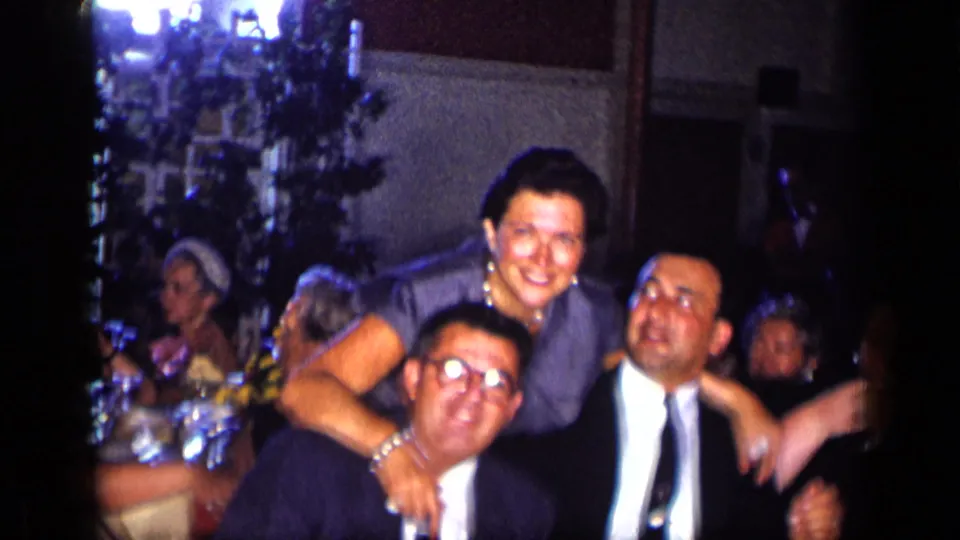 1957 New York home-movie frame of adults at a formal dinner table, including a woman in a blue dress leaning toward men in suits.
