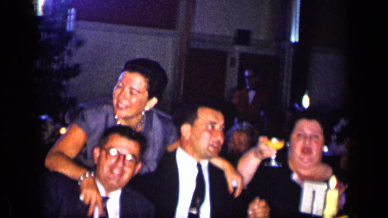 Woman in a blue dress laughs beside three men in suits, one holding a drink.