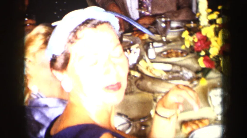 1957 New York home-movie frame of a formally dressed woman in a blue headscarf at a crowded dinner table.