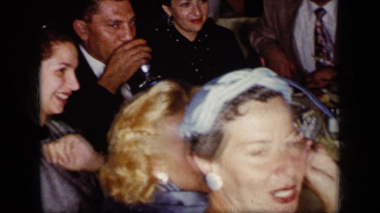 1957 New York dinner party guests sit around a table while a man drinks and others smile nearby.