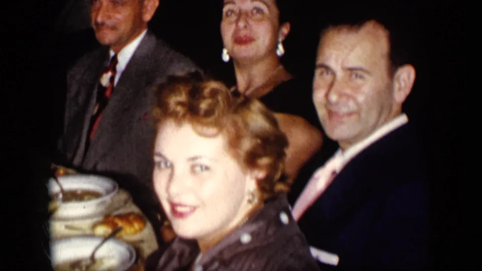 1957 New York dinner party frame with adults in suits and dresses looking toward the camera near table settings.