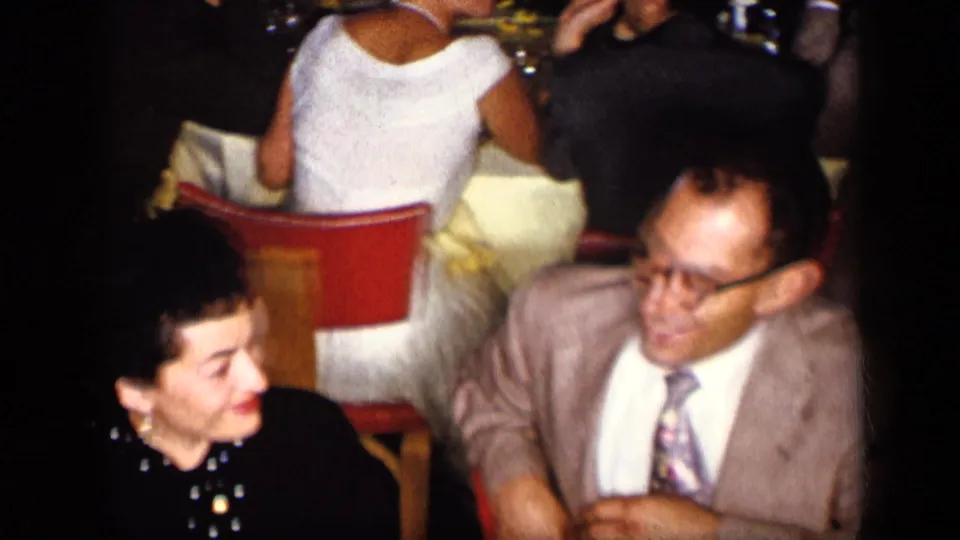 1957 New York home-movie frame of a woman in dark clothing and a man in a suit seated at a dinner event.