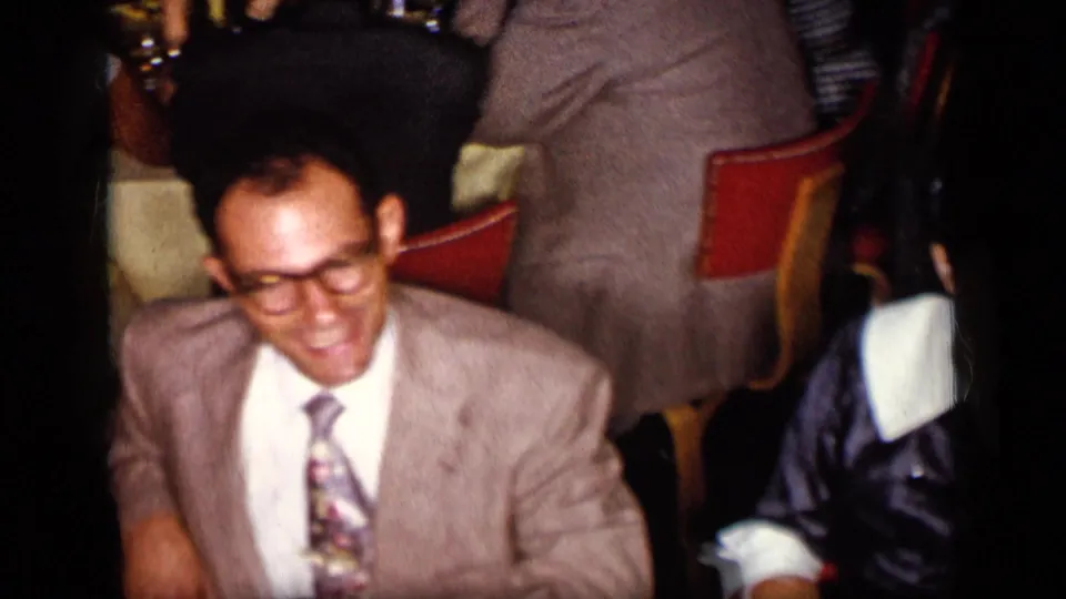 1957 New York home-movie frame with a smiling suited man at a crowded dinner gathering with red chairs nearby.