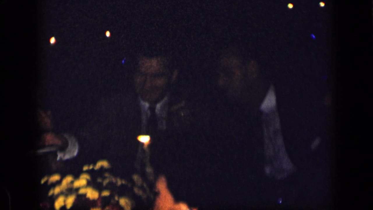 Suited men sit at a dim candlelit dinner table with flowers in front.