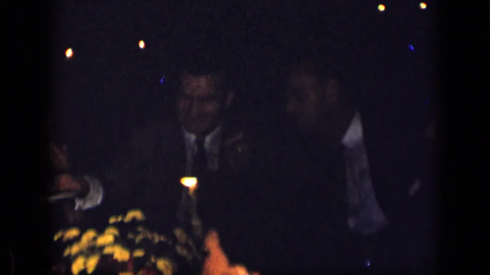 1957 New York home-movie frame showing suited dinner guests seated around a dark candlelit table with flowers.