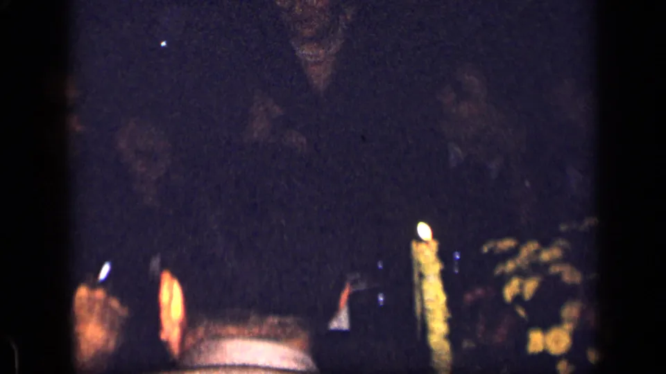Low-light 1957 New York home-movie frame showing partly obscured guests around a candlelit table.
