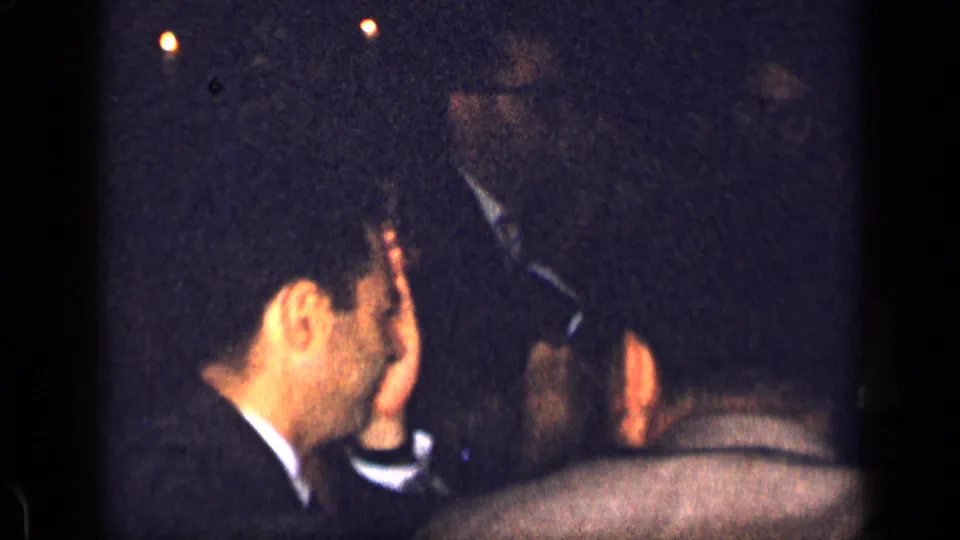 1957 New York home-movie frame showing suited men seated close together during a candlelit dinner party.