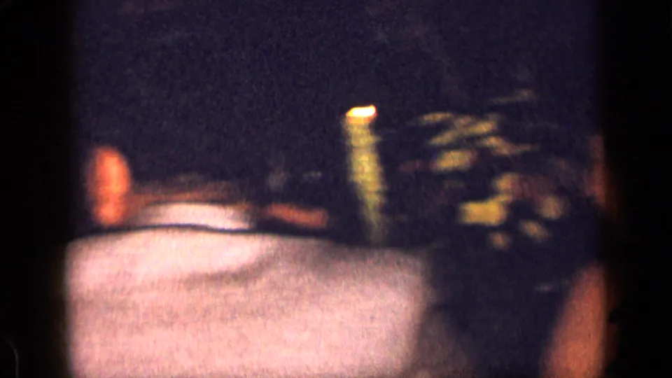 Dim 1957 New York home-movie frame with adults at a candlelit table, floral decor, and chair backs in the foreground.
