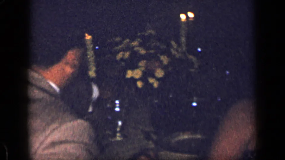 1957 New York 8mm frame showing a man in a light jacket near a candleholder, flowers, and glassware at a dark dinner table.