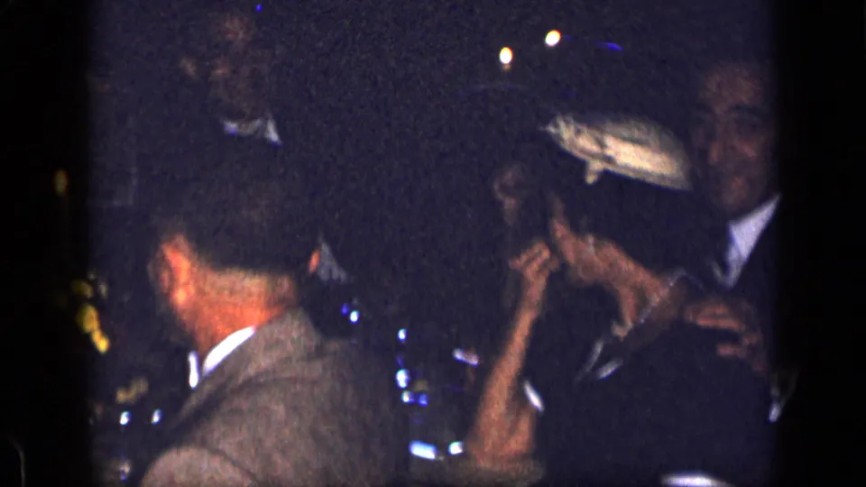 Candid 1957 New York home-movie frame of suited adults gathered closely around a low-light dinner table.