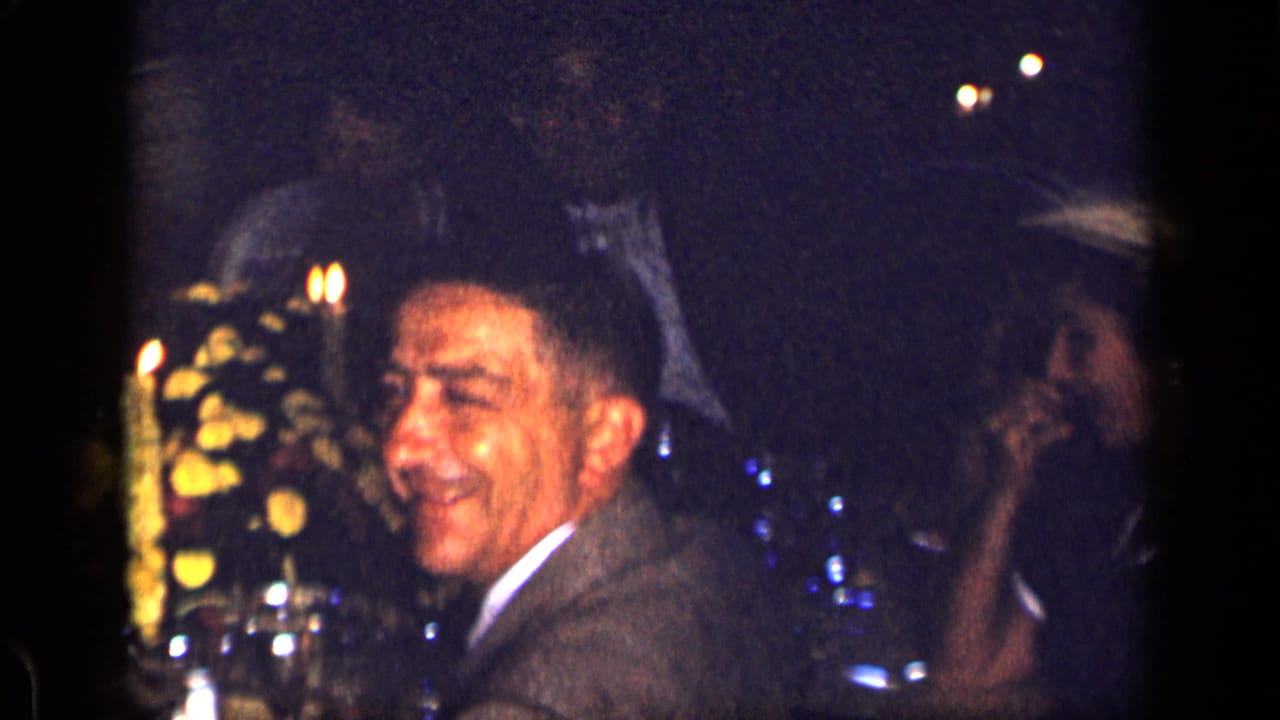 Man in a suit smiles at a candlelit restaurant table with flowers and glassware nearby.