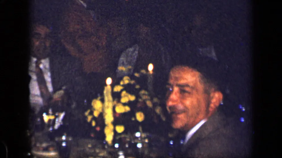 A suited diner turns toward the camera during a 1957 New York restaurant dinner, with candles and flowers on the table.