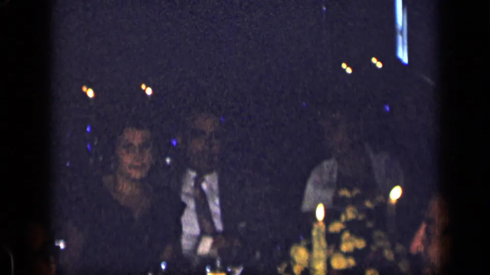 Several adults sit together at a candlelit restaurant table in 1957 New York, partly obscured by low light and film grain.