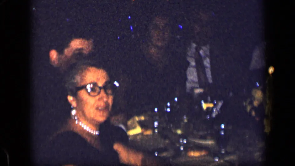 A woman wearing glasses and a pearl necklace sits at the 1957 New York restaurant dinner, with other guests and glassware nearby.