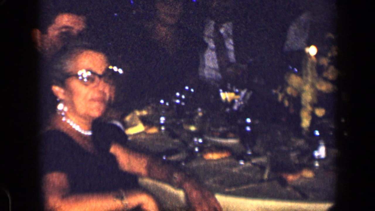 Woman in glasses and pearls seated at a dim dinner table with other guests nearby.