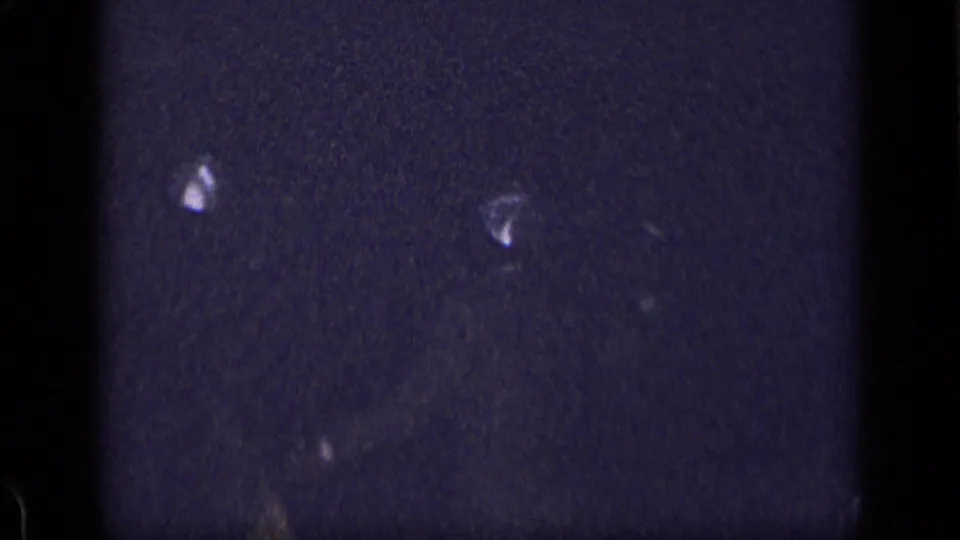 Dark 8mm home-movie frame with faint reflective shapes, heavy grain, and little identifiable subject detail.