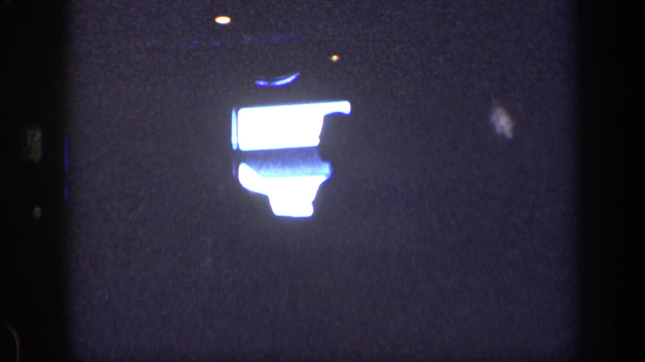 Dark frame with a bright glowing shape and faint silhouetted figure at right.